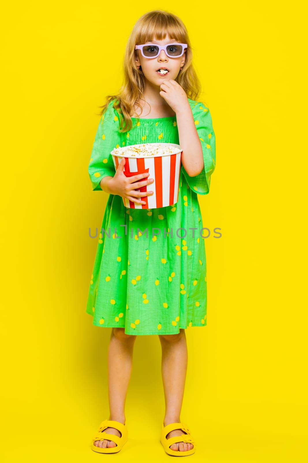 Excited young cute school girl in 3D glasses eating popcorn, watching interesting tv serial, sport game, film, online social media movie content. Teen female child kid on yellow background. Vertical
