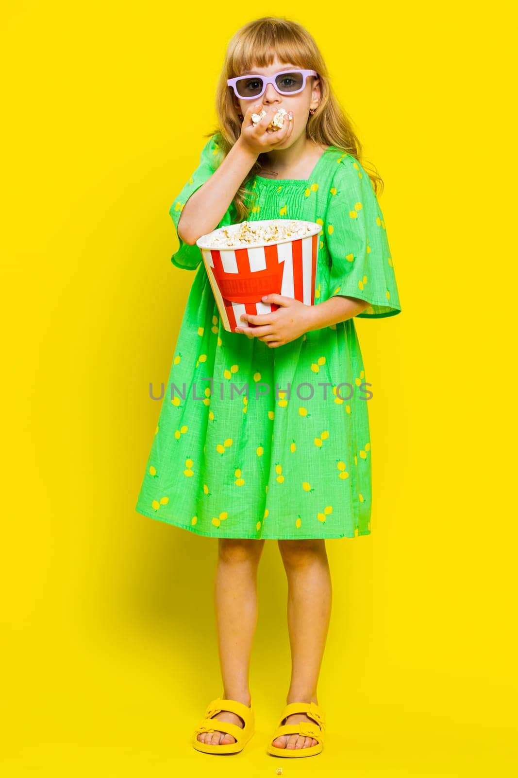 Excited young cute school girl in 3D glasses eating popcorn, watching interesting tv serial, sport game, film, online social media movie content. Teen female child kid on yellow background. Vertical