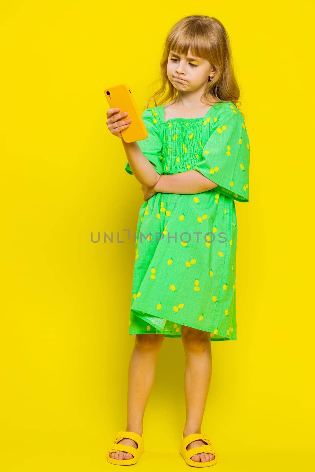 Unhappy blonde child girl kid use smartphone play online game, typing browsing, loses surprised sudden lottery results, bad fortune, loss, fail. Little preteen children on yellow background. Vertical