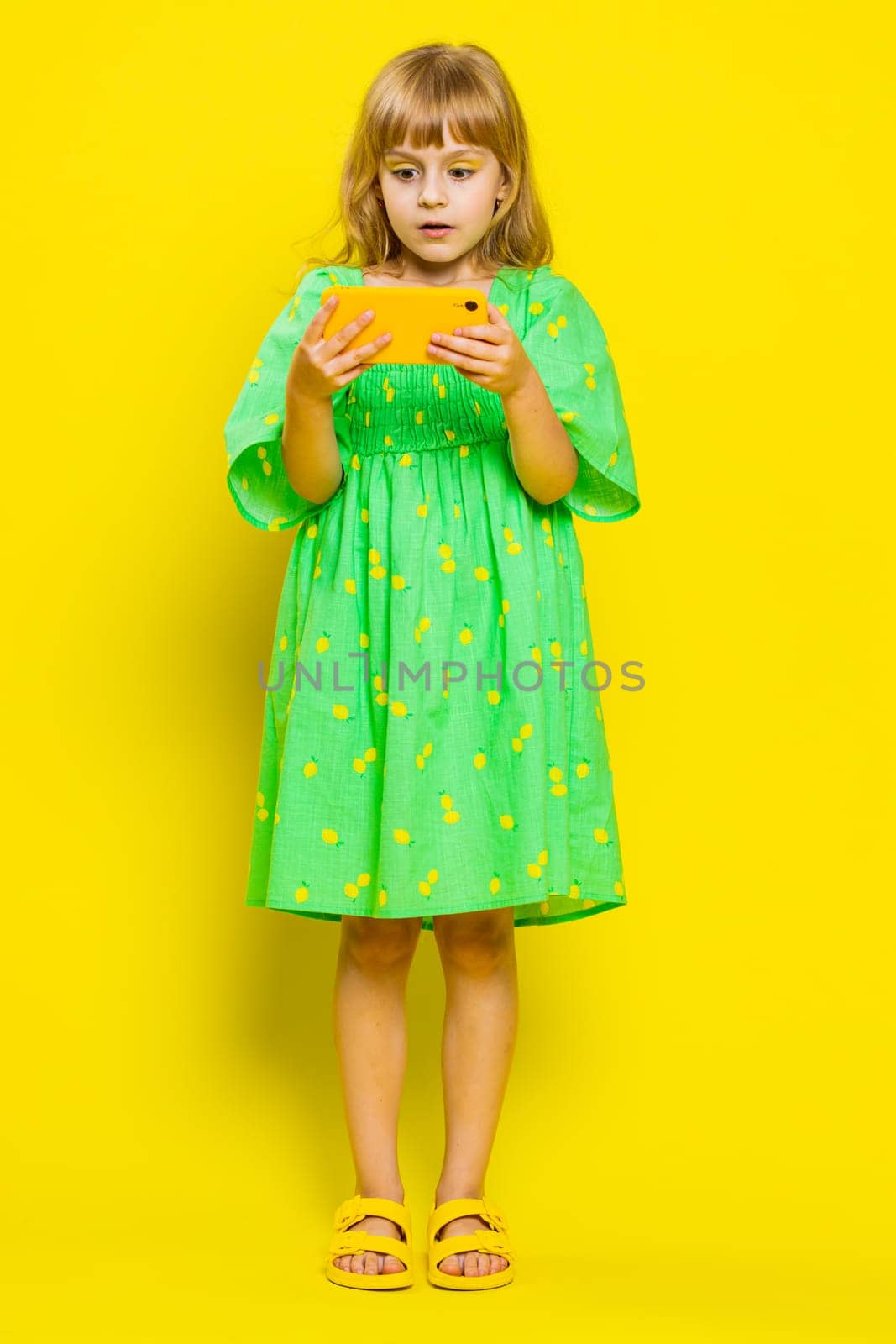 Worried funny young school girl enthusiastically playing racing or shooter video games on smartphone. Preteen child kid using smartphone gadget app with drive simulator on yellow background. Vertical