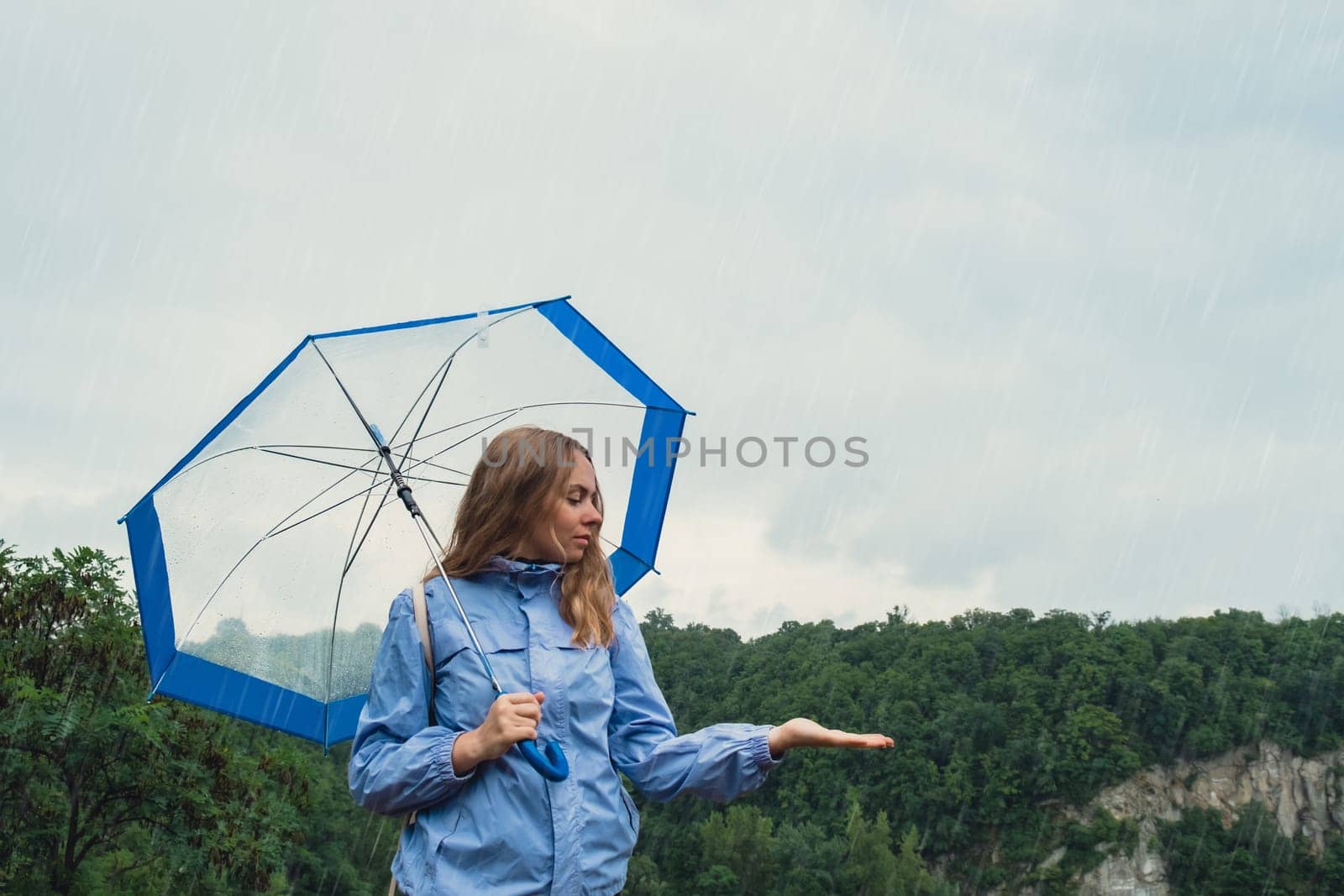 Blonde girl checking the weather under transparent umbrella outside in park. Autumn time rainy bad forecast conditions season. Tourist with umbrella travel in new places in rain. Landscape