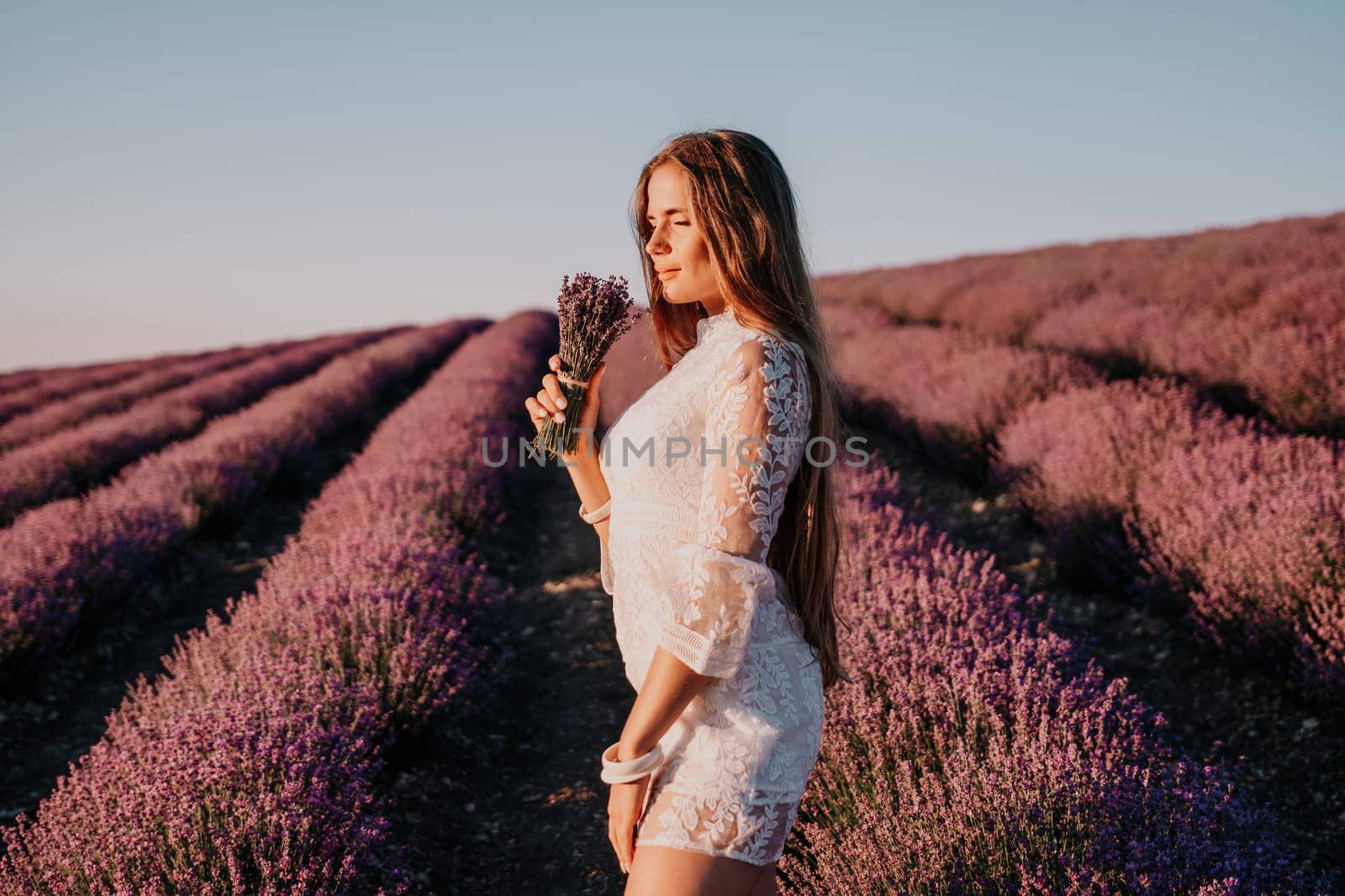 Close up portrait of young beautiful woman in a white dress and a hat is walking in the lavender field and smelling lavender bouquet.