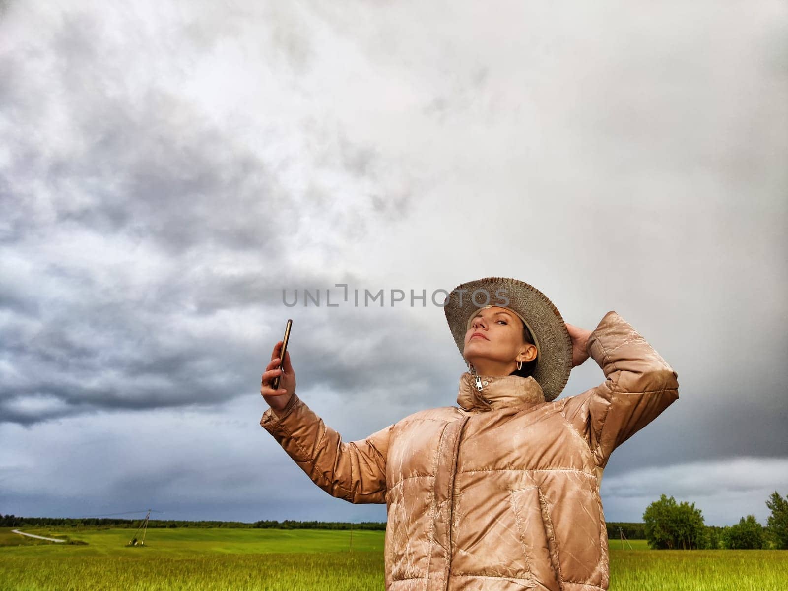 An adult girl in a field and with a stormy sky with clouds takes pictures of a rainbow and takes selfie in the rain. A woman having fun outdoors on rural and rustic nature