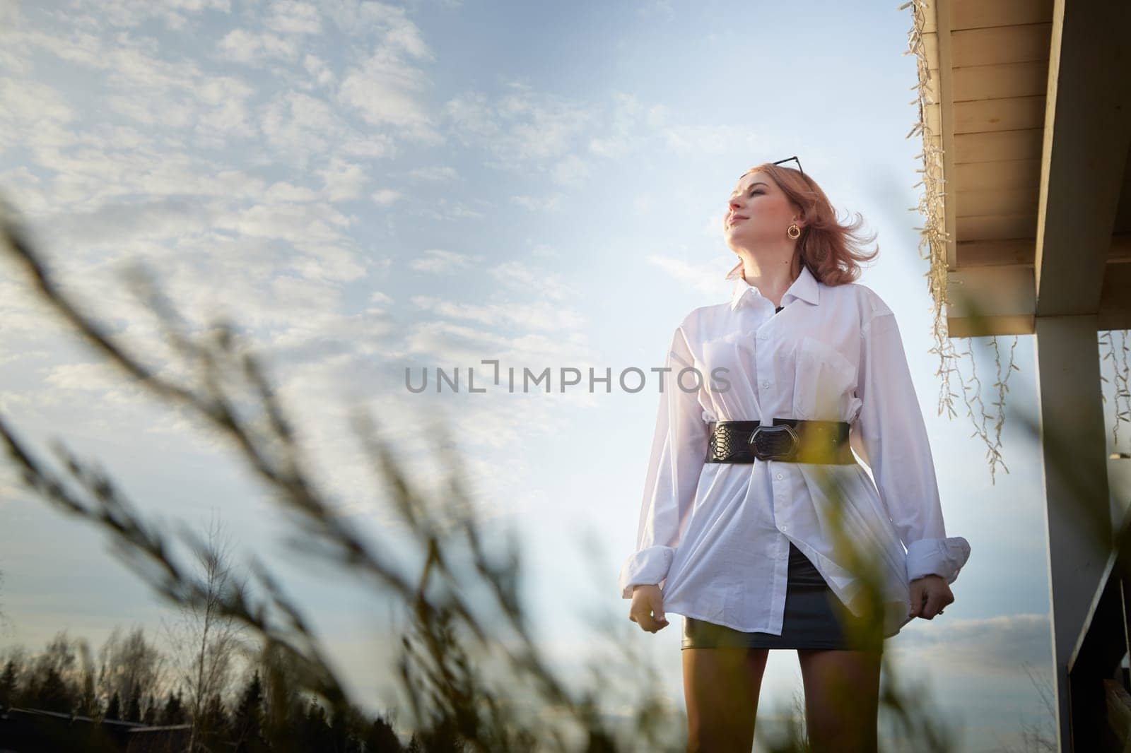 Beautiful girl with red hair in white shirt in open wooden pavillion in village or small town. Young slender woman and sky with clouds on background on autumn, spring or summer day