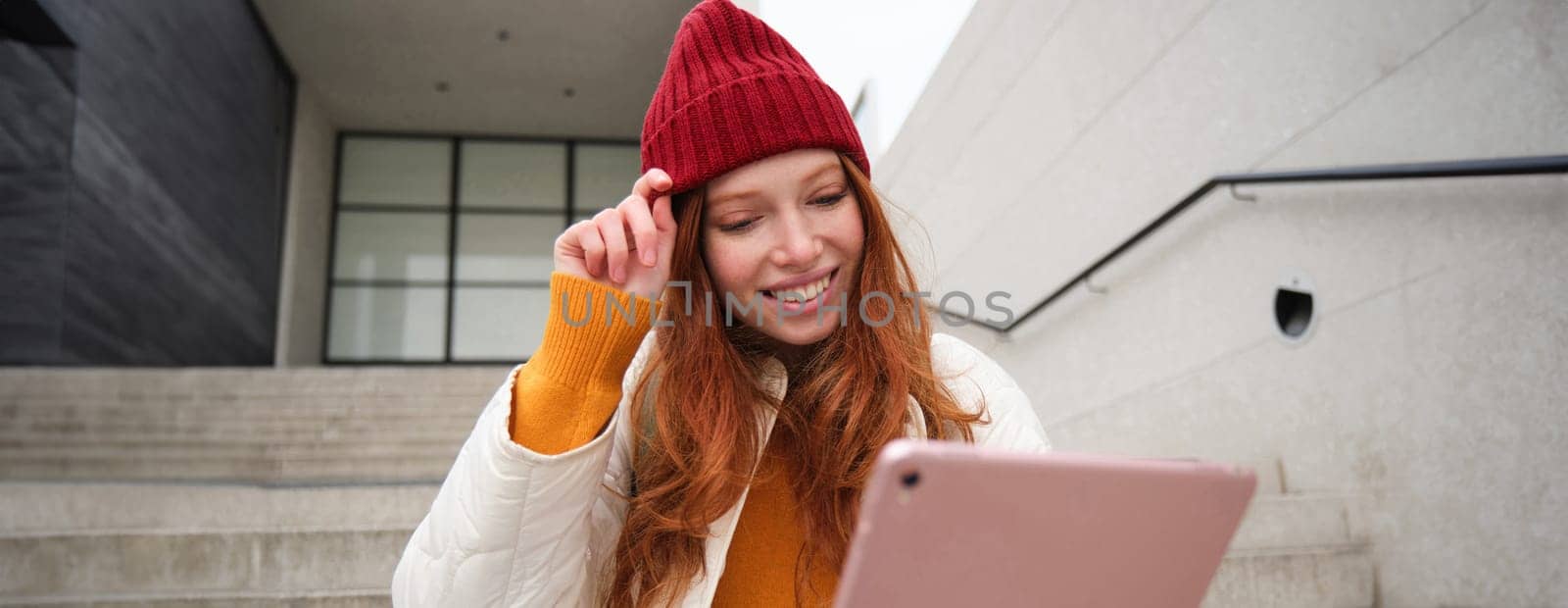 Beautiful redhead female model, ginger girl with digital tablet, sits on stairs outdoors, reads on her gadget, uses internet application and wifi on street.