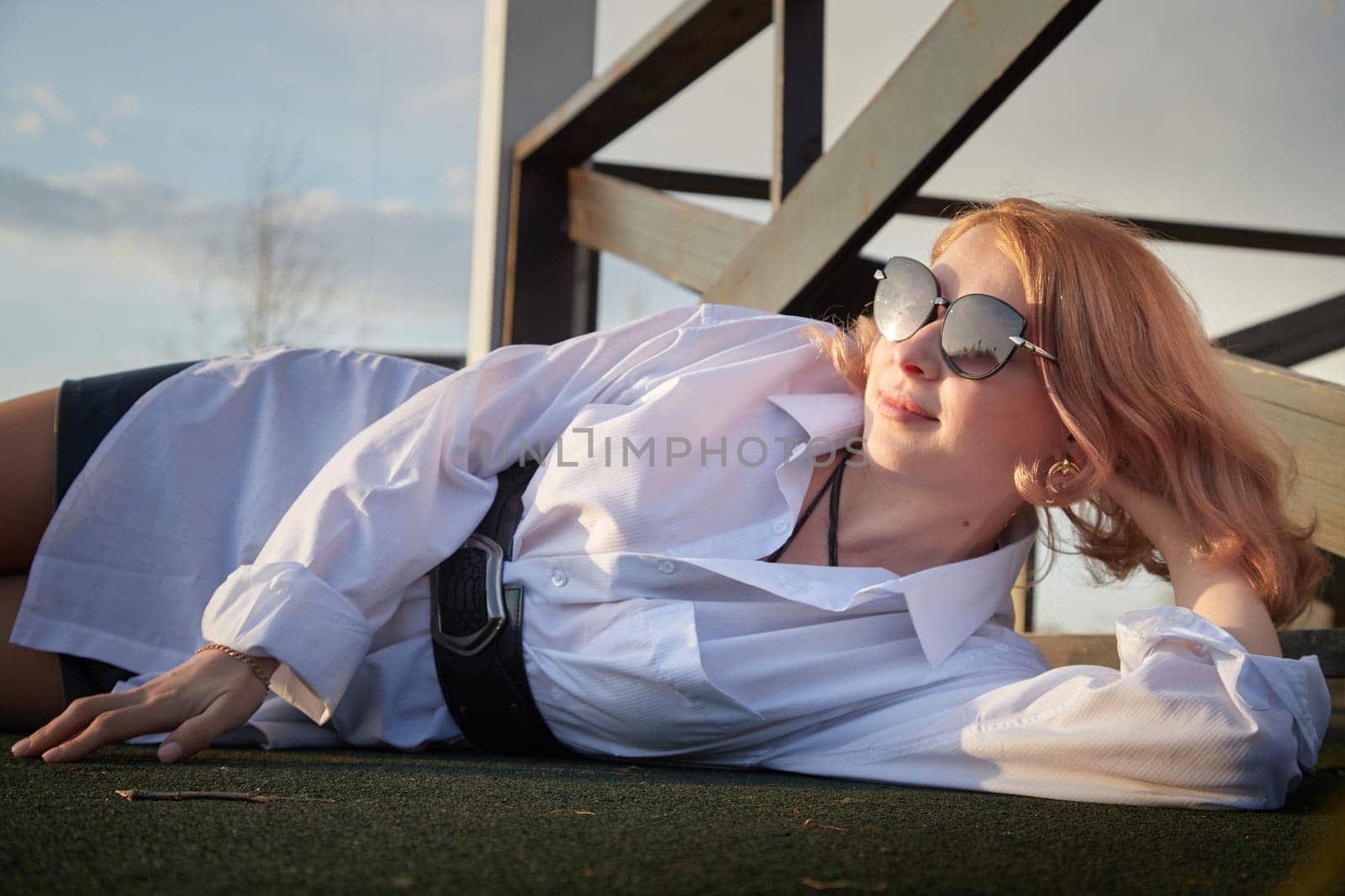 Beautiful girl with red hair in white shirt in open wooden pavillion in village or small town. Young slender woman and sky with clouds on background on autumn, spring or summer day