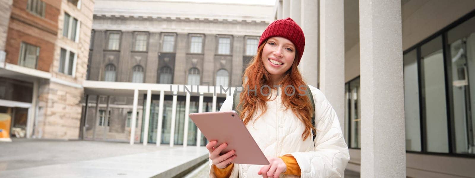 Happy redhead girl in red hat, walks around city with digital tablet, connects to public internet wifi and looks for route, looks at map on her gadget by Benzoix