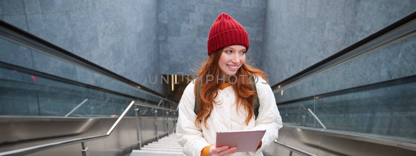 Beautiful redhead female model posing in city, walking up stairs with digital tablet, using gadget to plan her route, reading while going somewere.