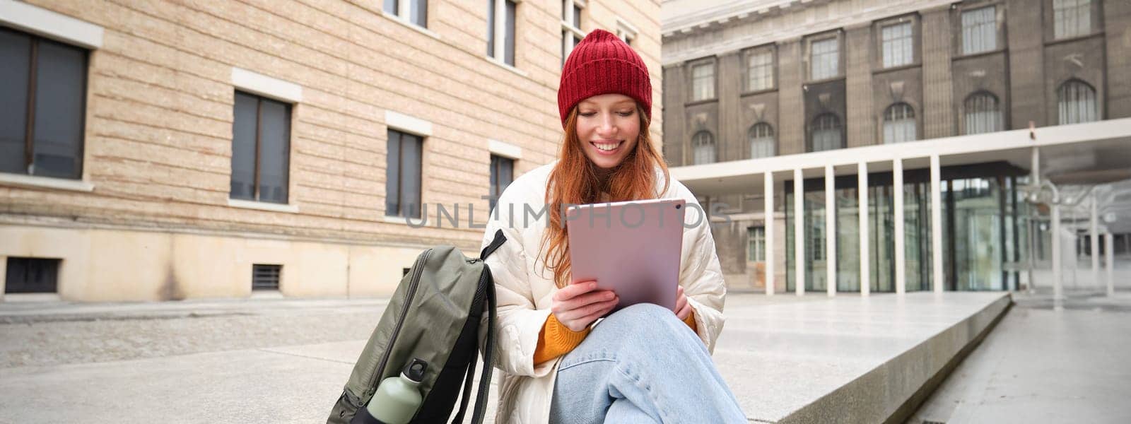 Redhead girl smiles, sits outdoors near building with digital tablet, thermos and backpack, connects to public internet and searches smth online on her gadget.