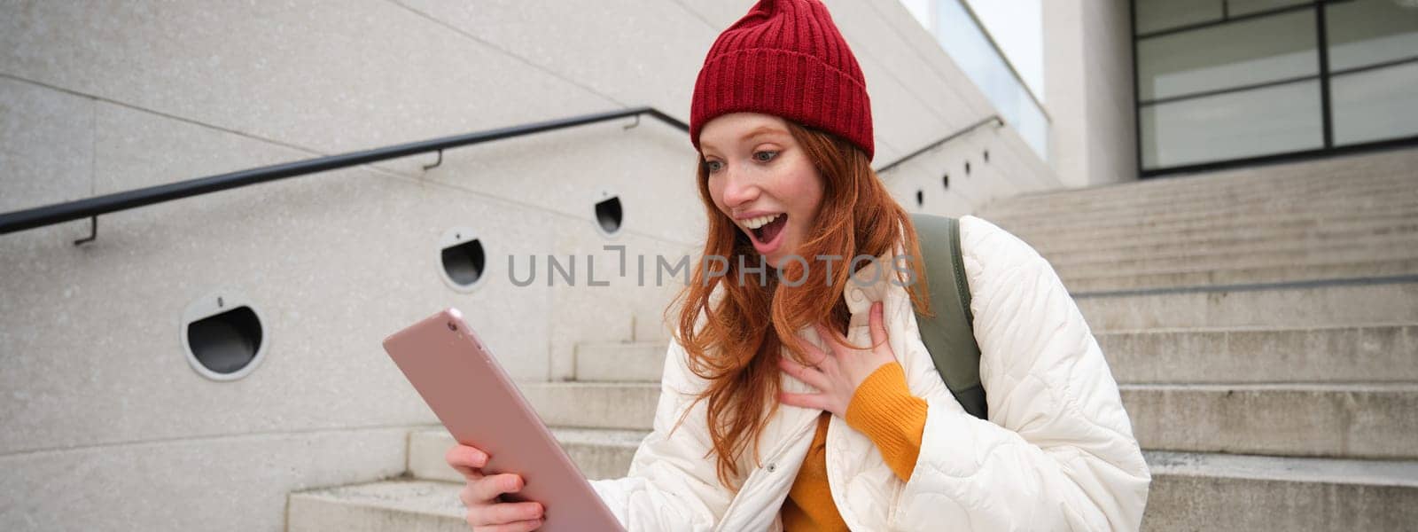 Happy stylish redhead girl, student in red hat, holds digital tablet, uses social media app, searches something online, connects to wifi.