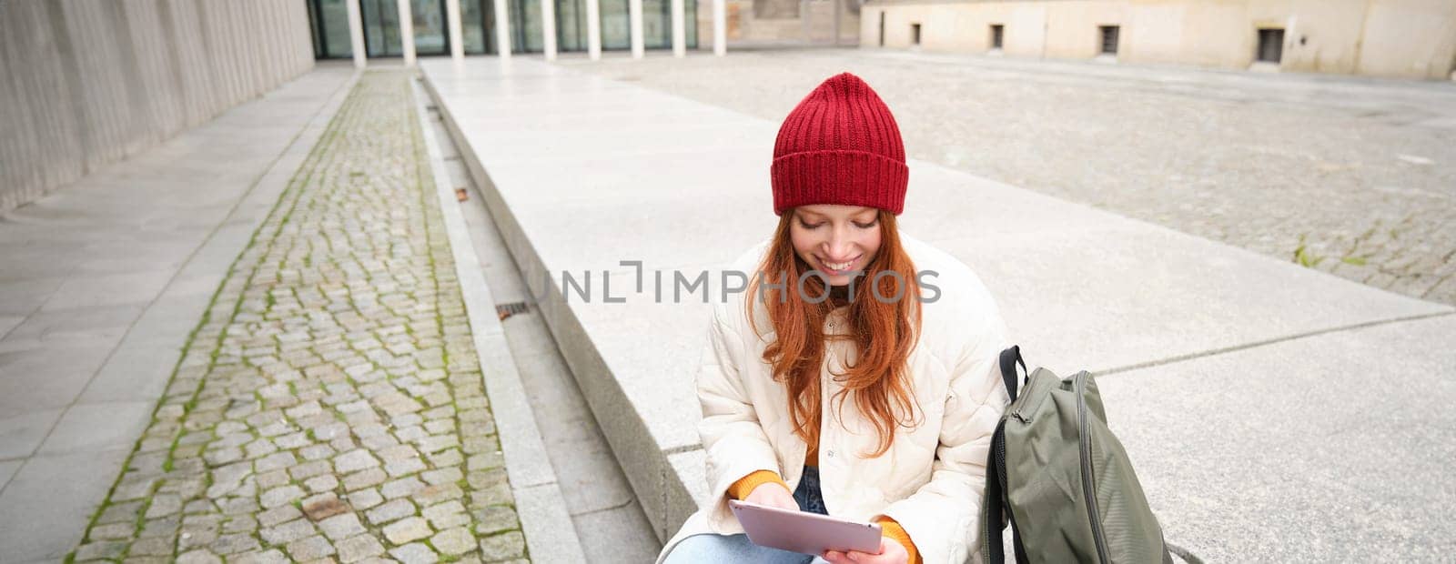 Beautiful redhead woman in red hat, sits with backpack and thermos, using digital tablet outdoors, connects to wifi, texts message, books tickets online by Benzoix