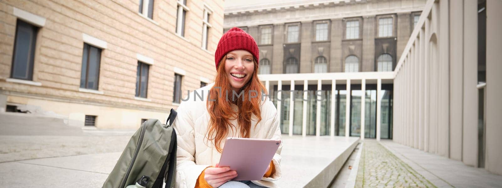 Beautiful redhead woman in red hat, sits with backpack and thermos, using digital tablet outdoors, connects to wifi, texts message, books tickets online by Benzoix