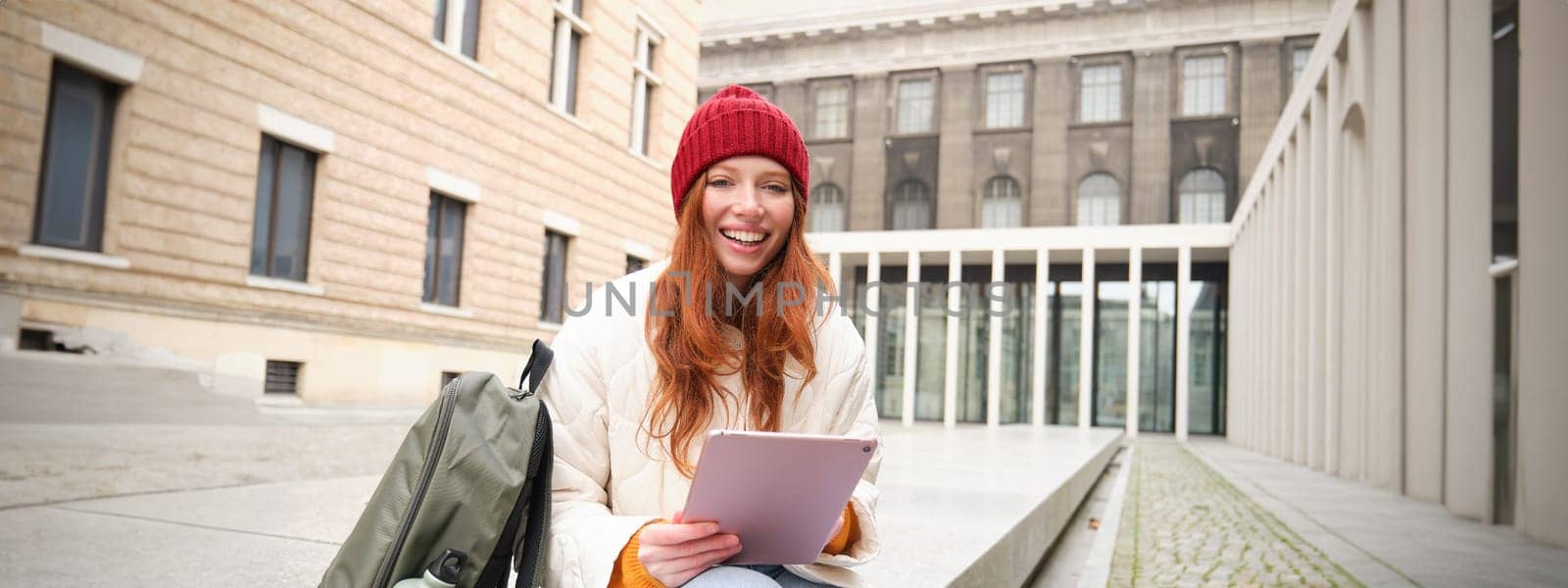 Beautiful redhead woman in red hat, sits with backpack and thermos, using digital tablet outdoors, connects to wifi, texts message, books tickets online.