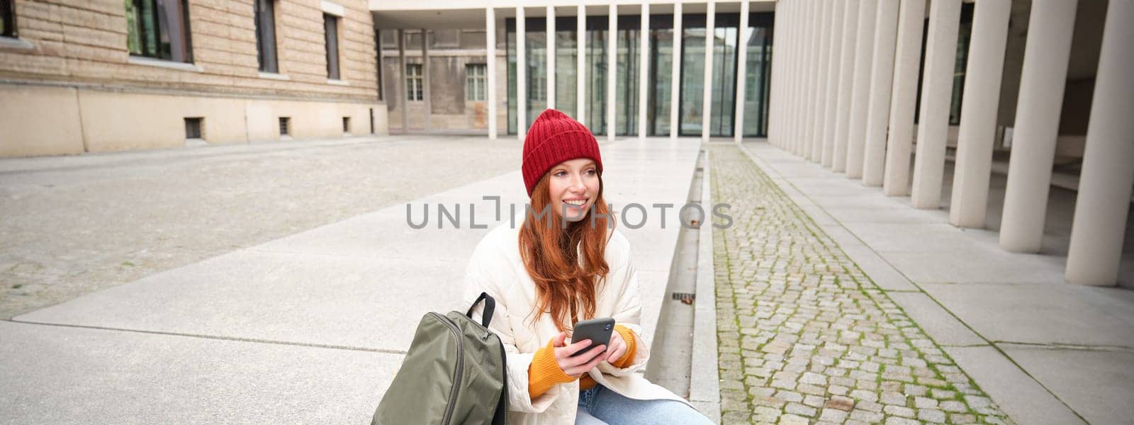 Social media and people. Young redhead girl sits on street, uses mobile phone app, looks up information in internet, holds smartphone.