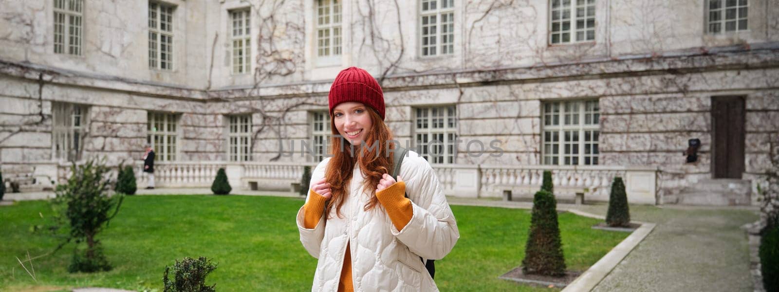 Portrait of smiling girl, backpacker in coat and red hat, wearing warm clothes for tourist trip around Europe in winter, walking around historical building.