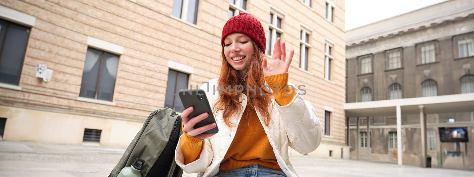 Social media and people. Young redhead girl sits on street, uses mobile phone app, looks up information in internet, holds smartphone by Benzoix