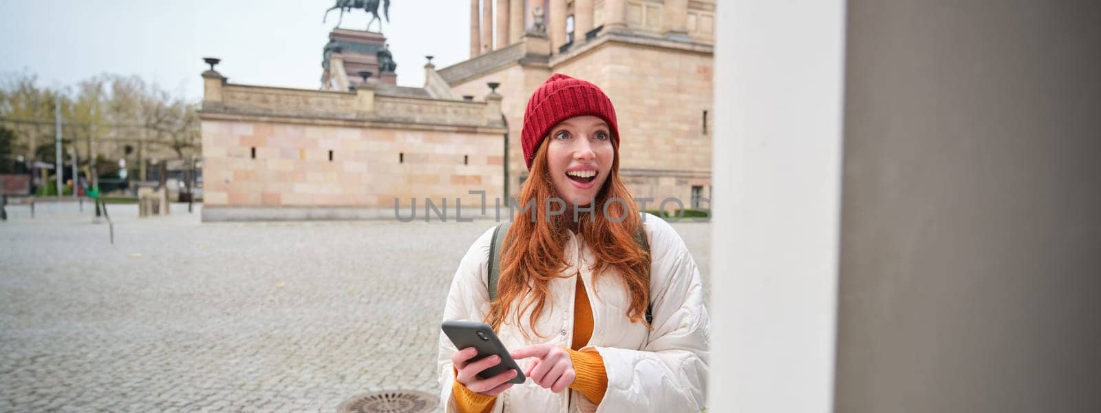 Tourism and sightseeing concept. Young redhead woman, tourist walks around city, looks at her smartphone app and at history stand, explores adventures.