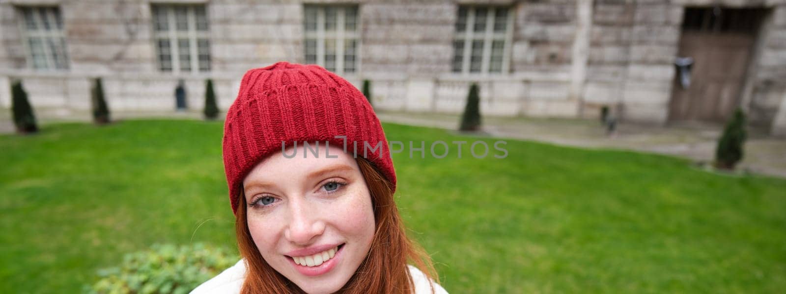 Vertical portrait of cute redhead female student in red hat and warm gloves, sits in park on bench, smiles and looks cute at camera.