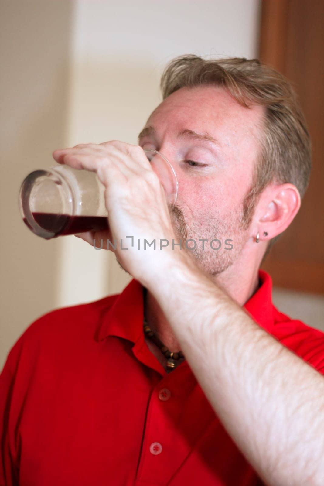 Middle aged man with his arm raised drinking a dark purple fruit juice out of a tall clear beverage glass.