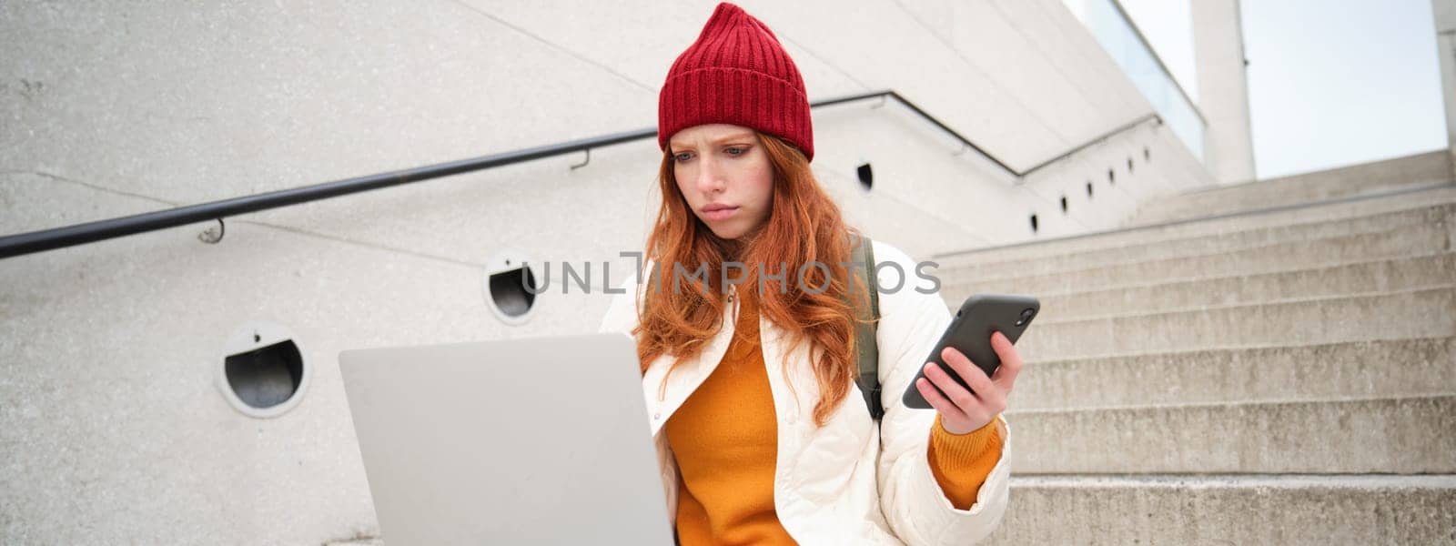 Portrait of redhead woman sits on stairs, uses laptop and holds smartphone, looks confused and upset at computer screen, tries to connect to wifi public internet by Benzoix