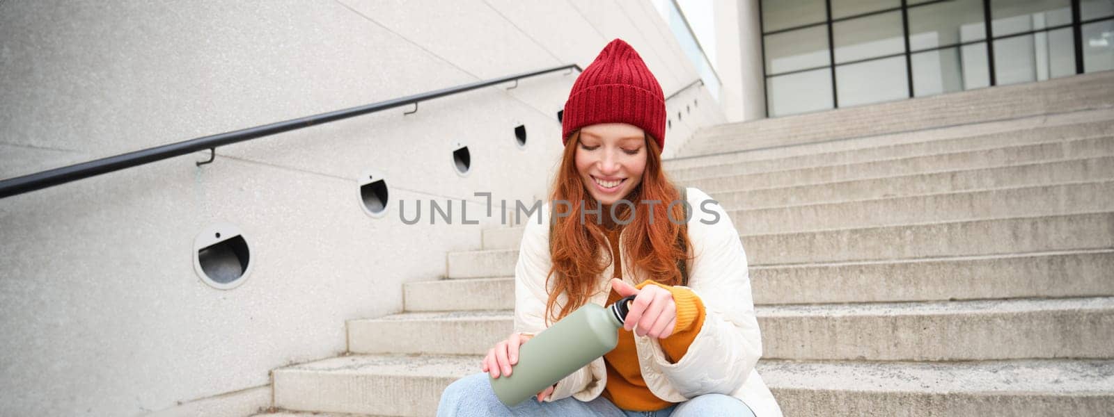 Happy redhead woman, tourist with thermos, drinks her hot tea, coffee from travel flask, restests during her travelling in city and sightseeing by Benzoix