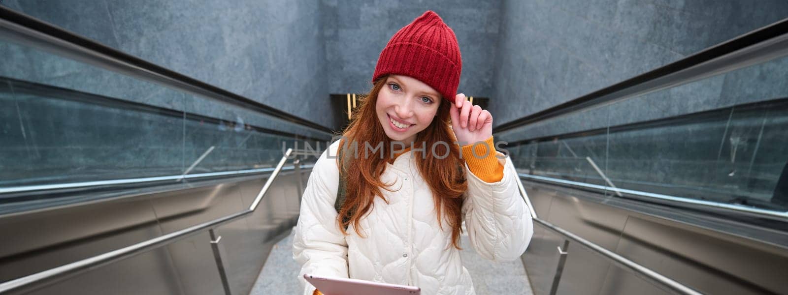 Smiling redhead woman walking up stairs with digital tablet, using gadget application while going somewhere in city.