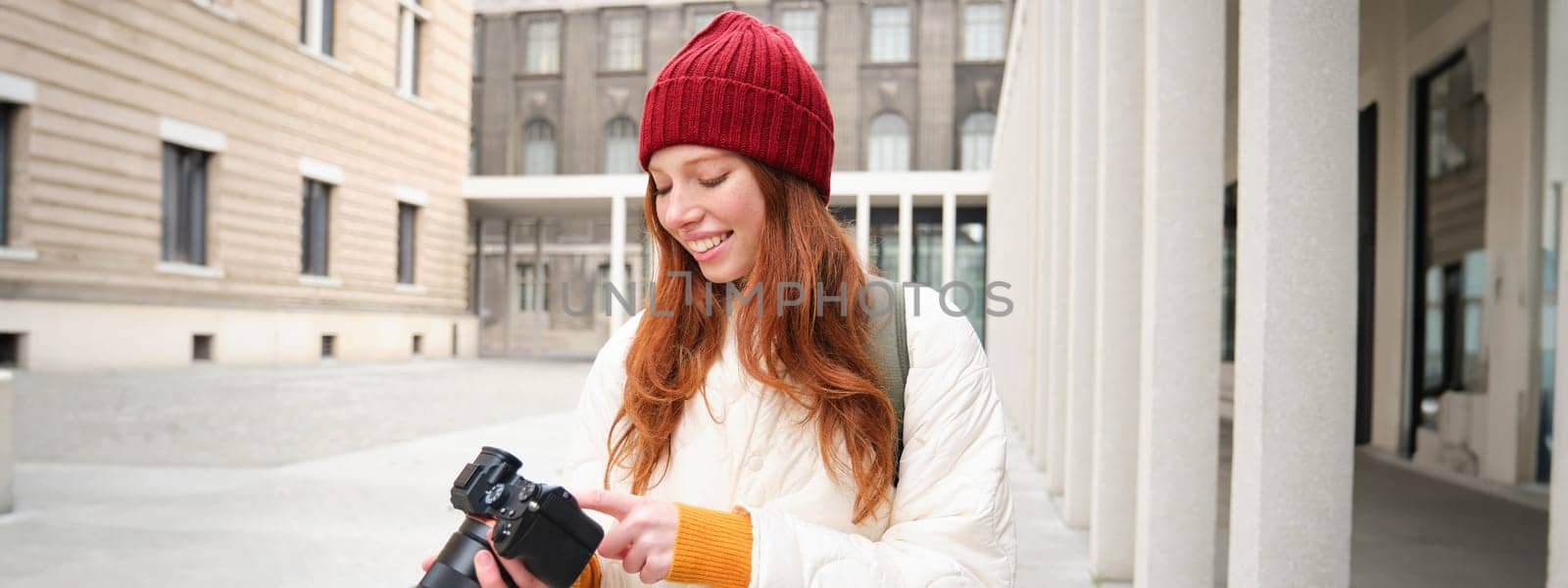 Smiling redhead girl photographer, taking pictures in city, makes photos outdoors on professional camera. Young talent and hobby concept