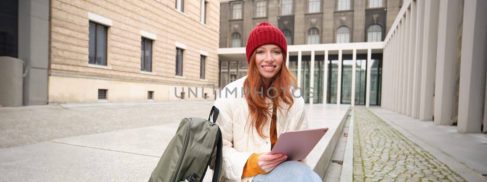 Beautiful redhead woman in red hat, sits with backpack and thermos, using digital tablet outdoors, connects to wifi, texts message, books tickets online.