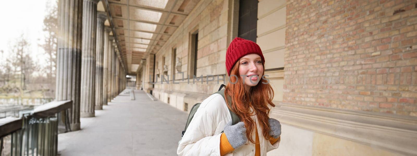 Smiling redhead girl with backpack, walks in city and does sightseeing, explore popular landmarks on her tourist journey around Europe by Benzoix