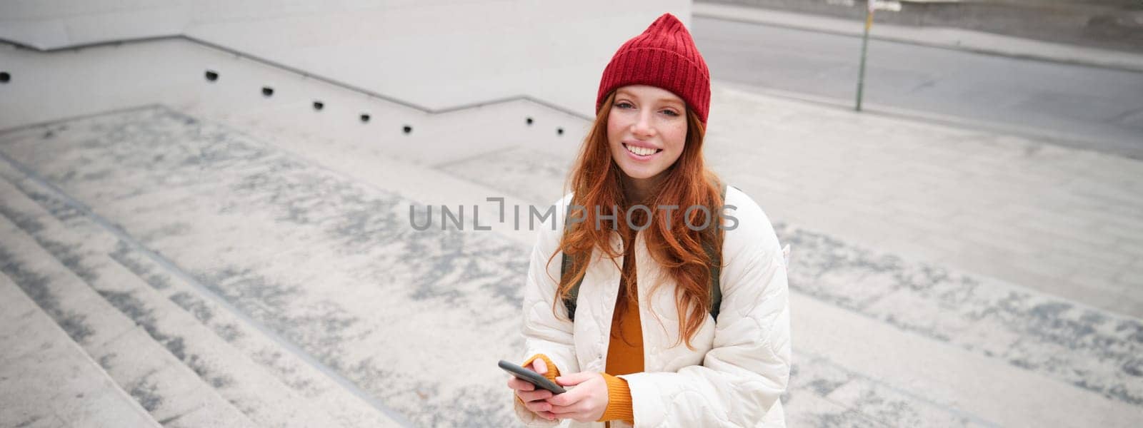 Smiling redhead girl, student tourist walks around city, goes up the stairs, looks at mobile phone map to get around town, sends message on smartphone.