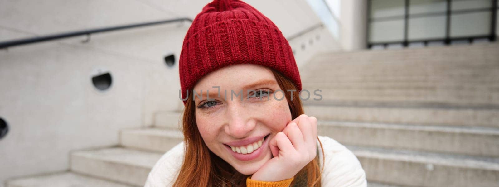 Beautiful redhead student, girl in red hat, smiles sincere, looks happy and relaxed, sits on stairs outdoors. Copy space