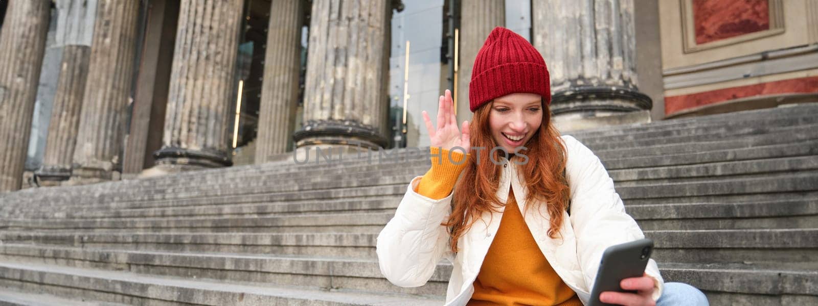 Young redhead woman sits on stairs outdoors and waves hand at smartphone camera, video chats with friends, connects to public wifi.