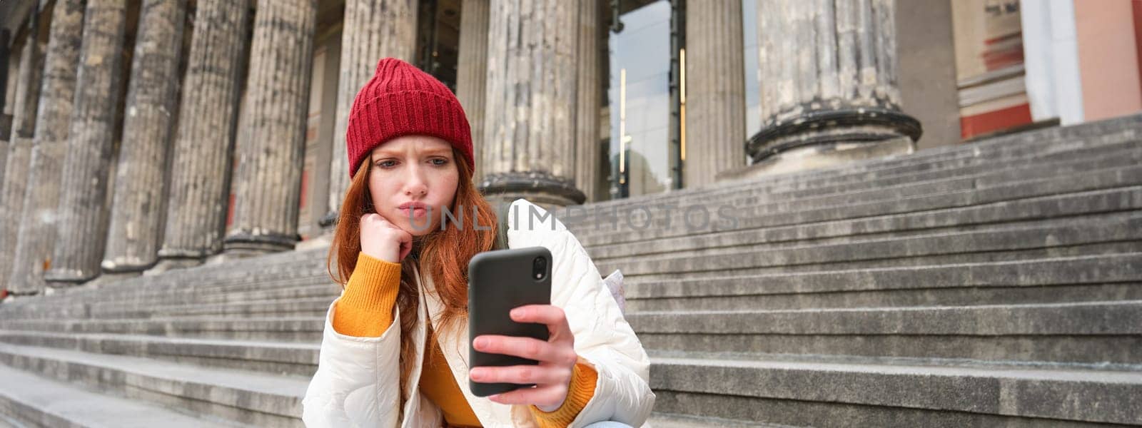 Portrait of young redhead woman with complicated face, sits on street stairs in red hat, holds smartphone and frowns thoughtful, feels uneasy.