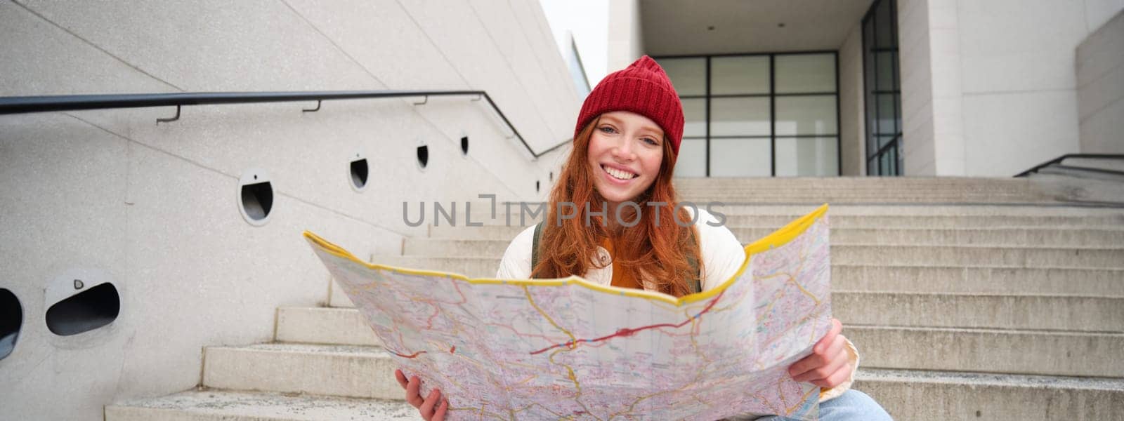 Young smiling redhead girl, tourist sits on stairs outdoors with city paper map, looking for direction, traveller backpacker explores city and looks for sightseeing by Benzoix