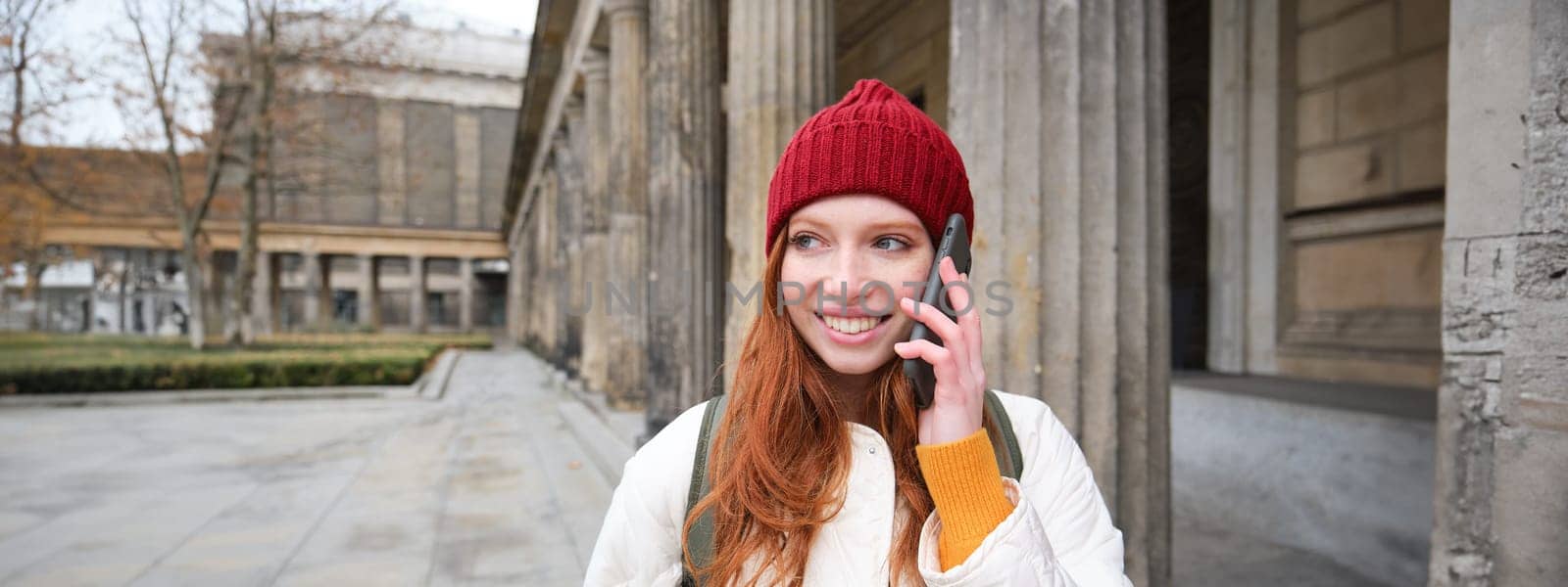 Smiling redhead female tourist talks on mobile phone and walks around city. Happy student in red hat calls friend, stands on street and uses smartphone by Benzoix