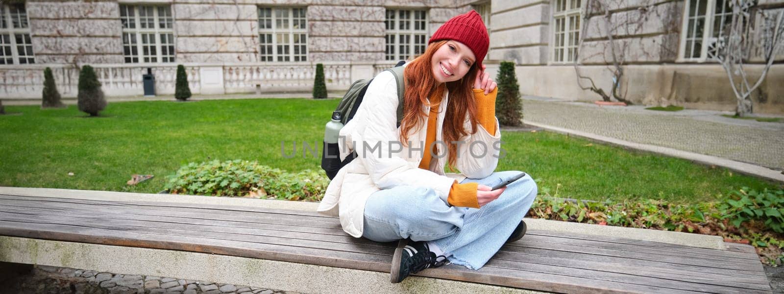 Portrait of stylish young woman, 25 years, sits on bench in park and uses mobile phone, reads online news, messages or watches video on smartphone app, connects to public wifi.