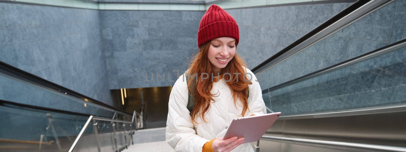 Smiling redhead woman walking up stairs with digital tablet, using gadget application while going somewhere in city by Benzoix