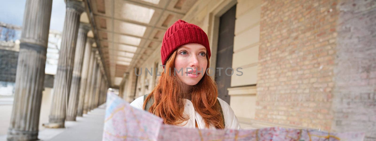 Adventurous redhead girl walks in town with paper map, explores city as tourist, looks for popular tourism attractions, looks around excited and smiles by Benzoix