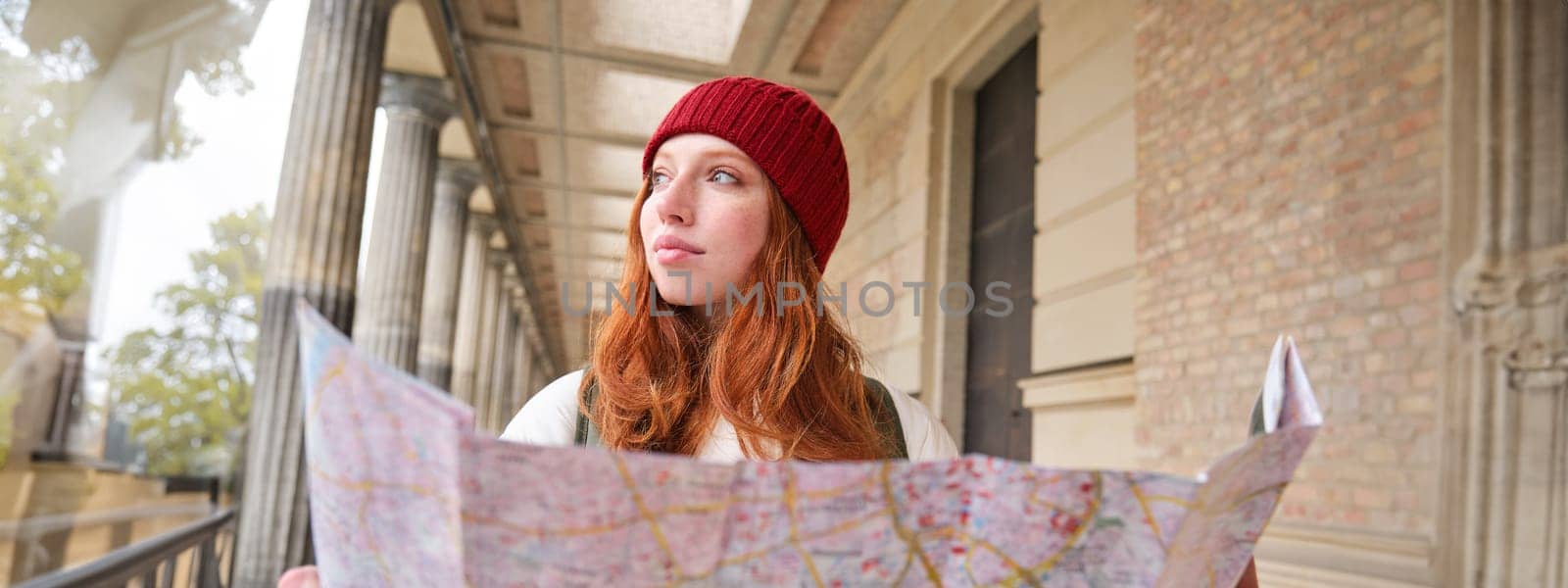 Smiling young redhead woman in red hat, looks at paper map to look for tourist attraction. Tourism and people concept. Girl explores city, tried to find way by Benzoix