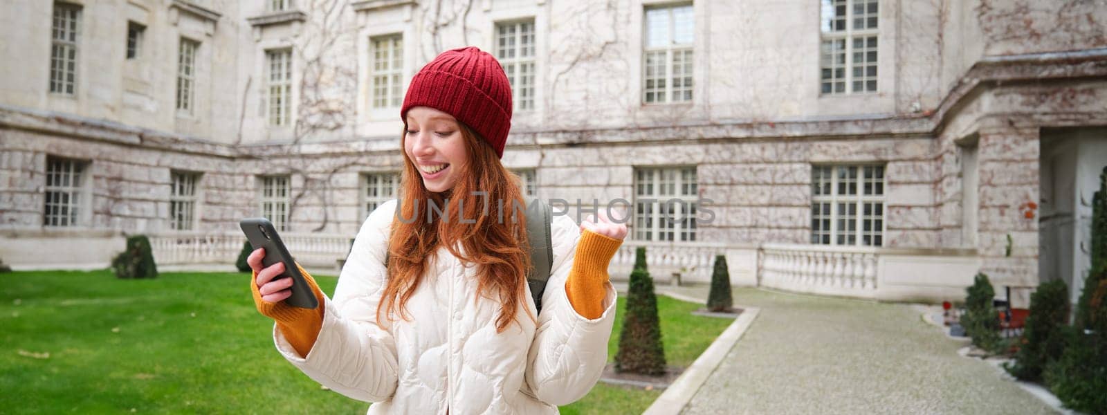 Cheerful redhead girl winning, celebrating victory, read great news on mobile phone and jumping from joy, saying yes, standing outside building in yard by Benzoix