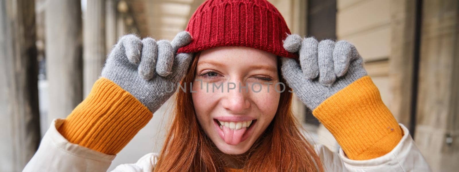 Portrait of young redhead woman in knitted hat and gloves, smiles and looks aside, walks around city in winter. Copy space