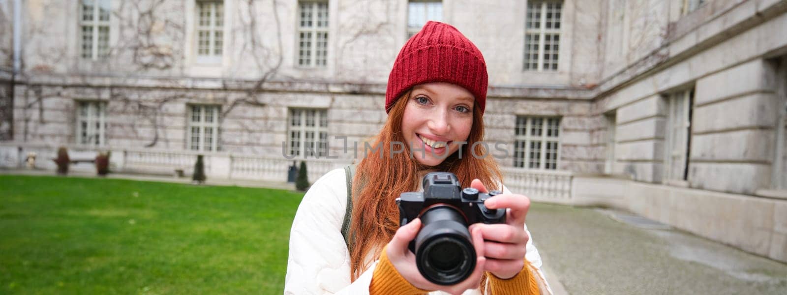 Redhead girl photographer takes photos on professional camera outdoors, captures streetstyle shots, looks excited while taking pictures.