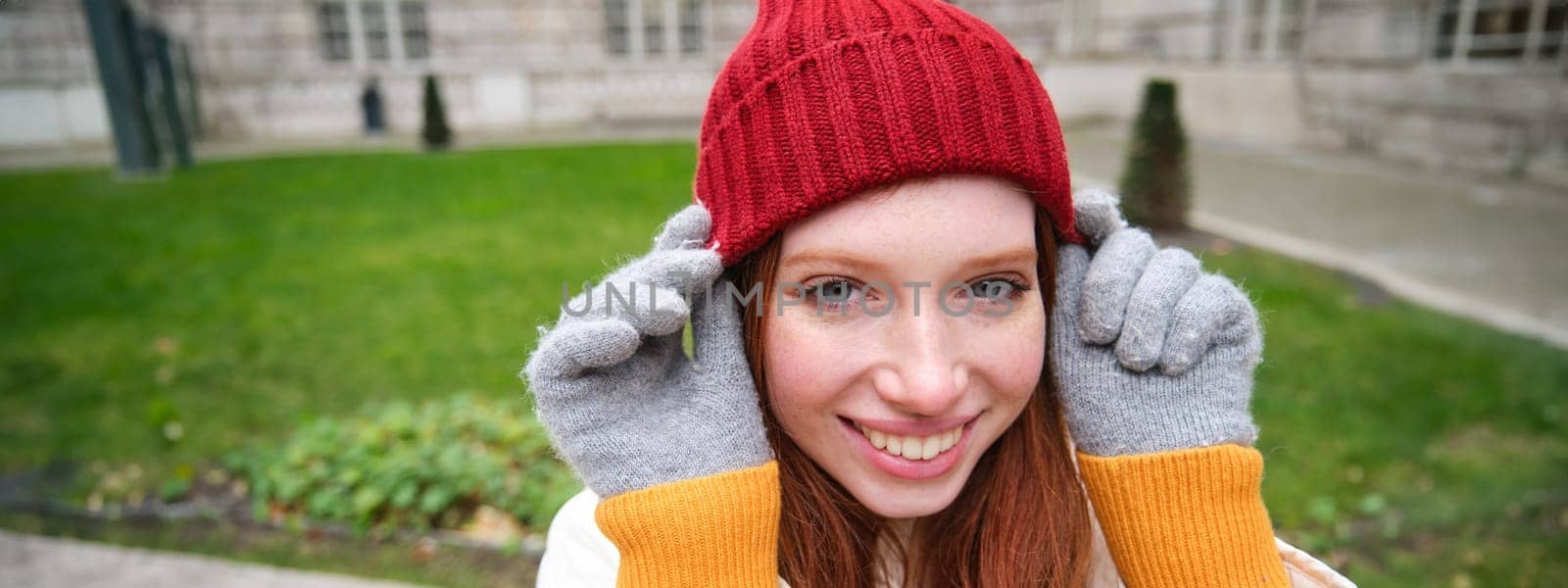 Close up portrait of beautiful redhead woman in red knitted hat, warm gloves, smiling and looking happy at camera, sitting in park.