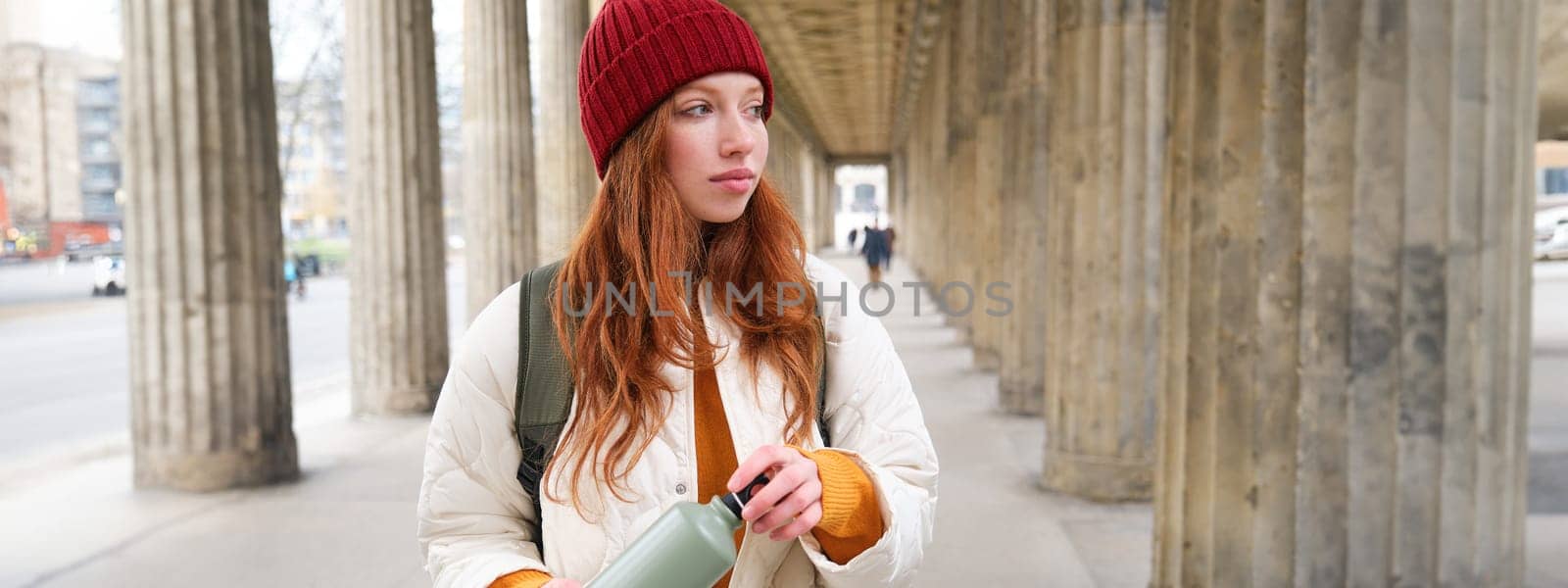 Young redhead girl holds thermos in hands, pours herself a hot drink while walking in city. Tourist takes break, opens flask for refreshment beverage.