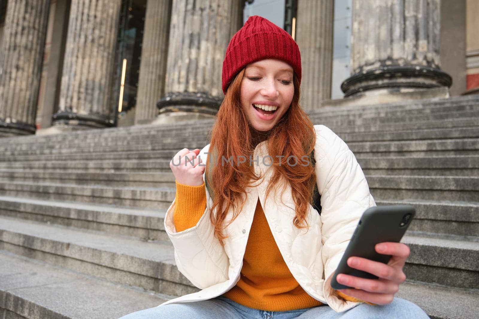 Achievement celebration. Happy redhead girl sits on stairs outdoors and looks at phone, triumphs, wins something and looks satisfied at mobile screen.