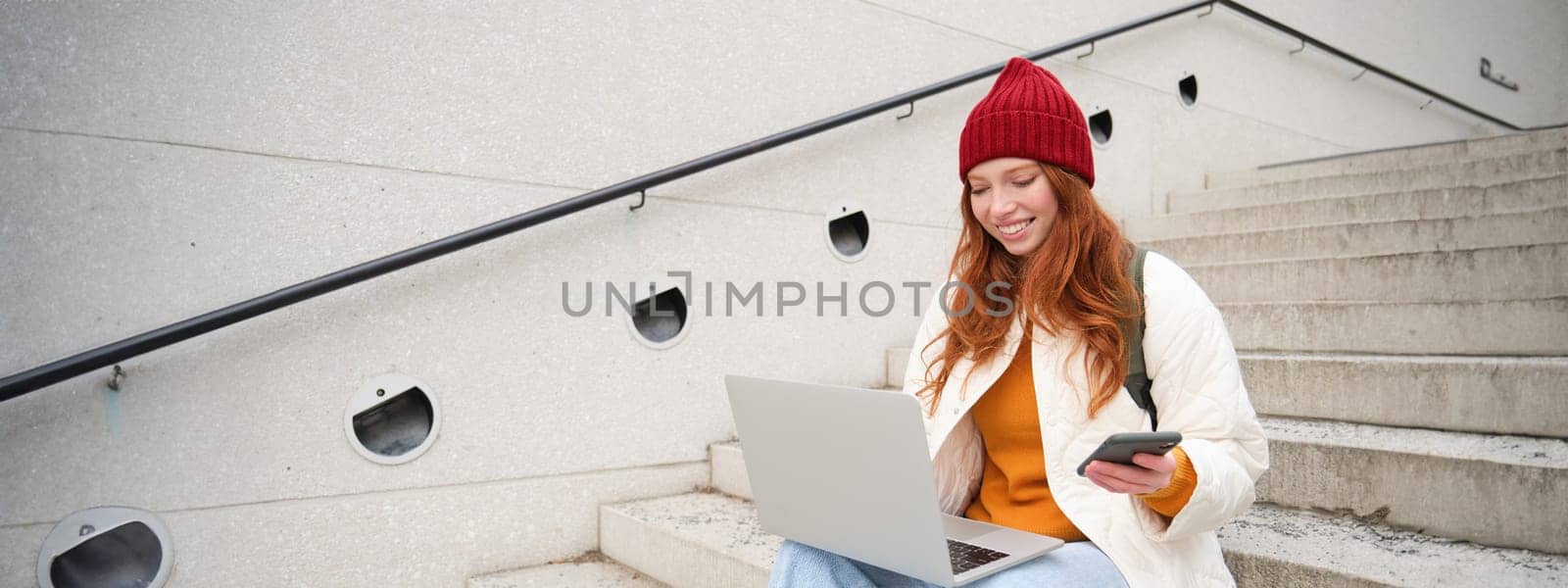 Young tourist, girl booking hotel room on laptop, enters confirmation code on smartphone, sitting with computer and phone on stairs outdoors by Benzoix