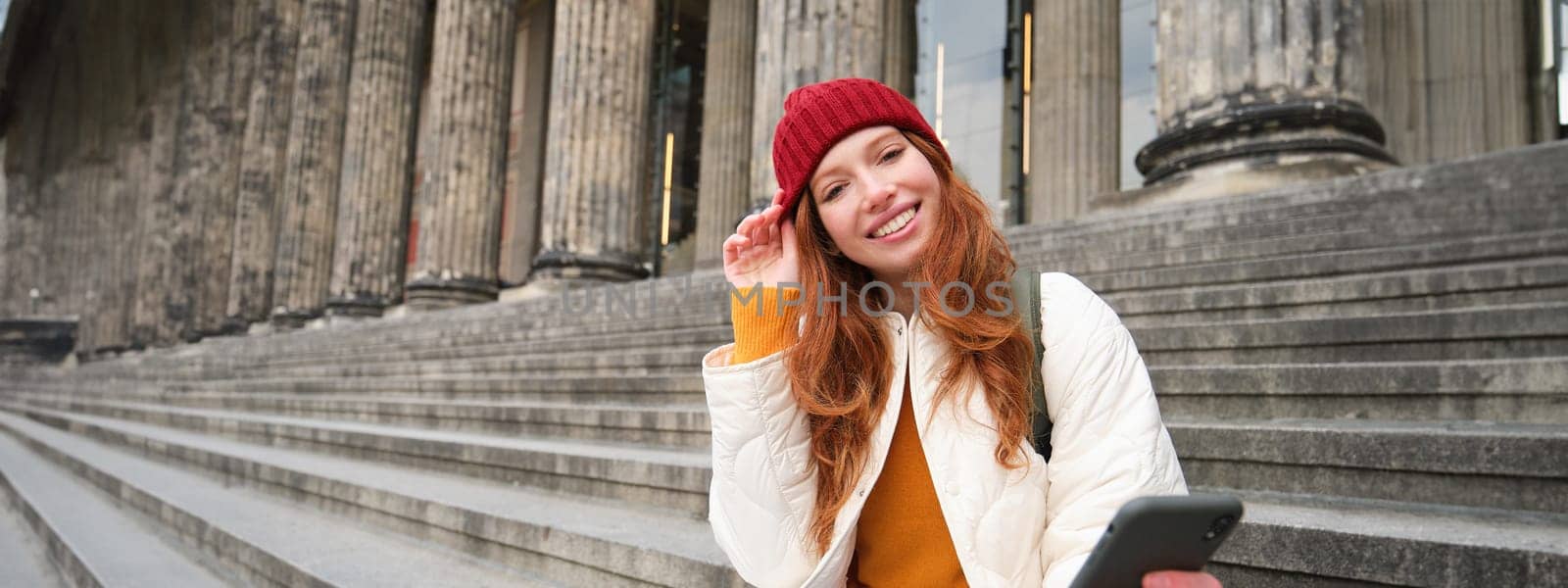 Portrait of young redhead woman in red hat, sitting on stairs, tourist looks at her mobile phone, rests on staircase of museum, connects public wifi by Benzoix