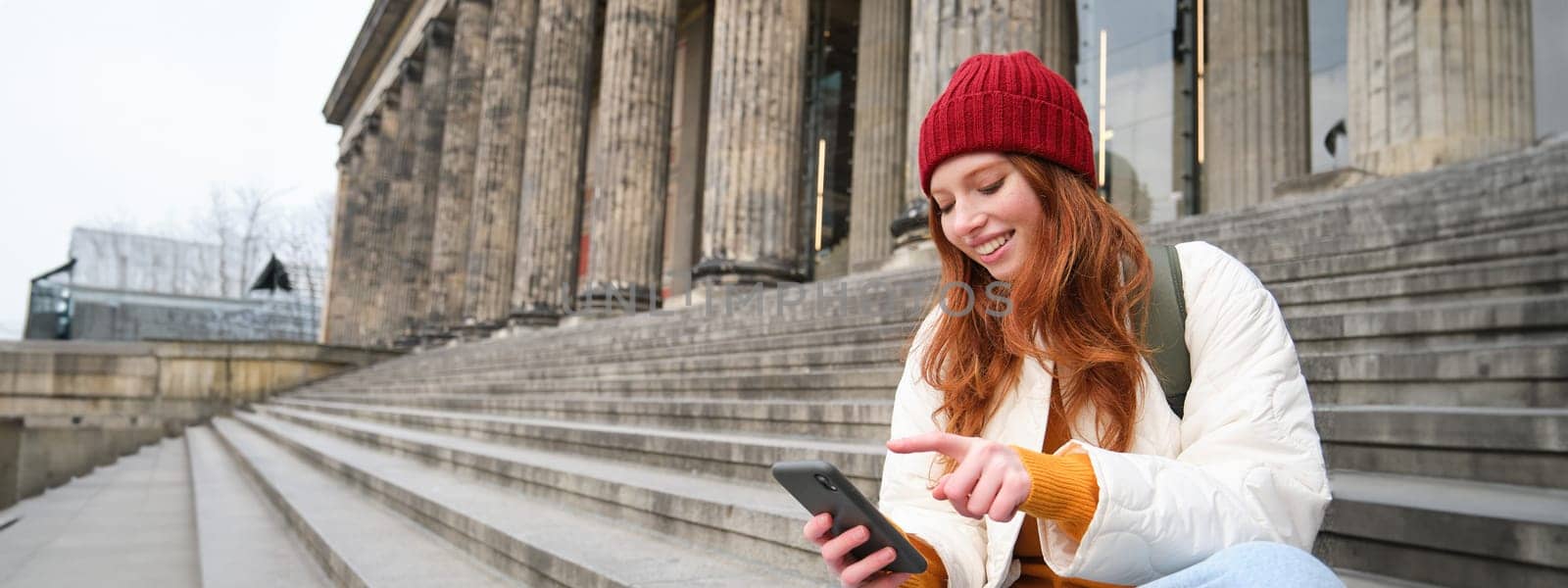 Stylish young redhead woman, talking on mobile phone app, using social media application, looking for something online on smartphone, sits on stairs outdoors.