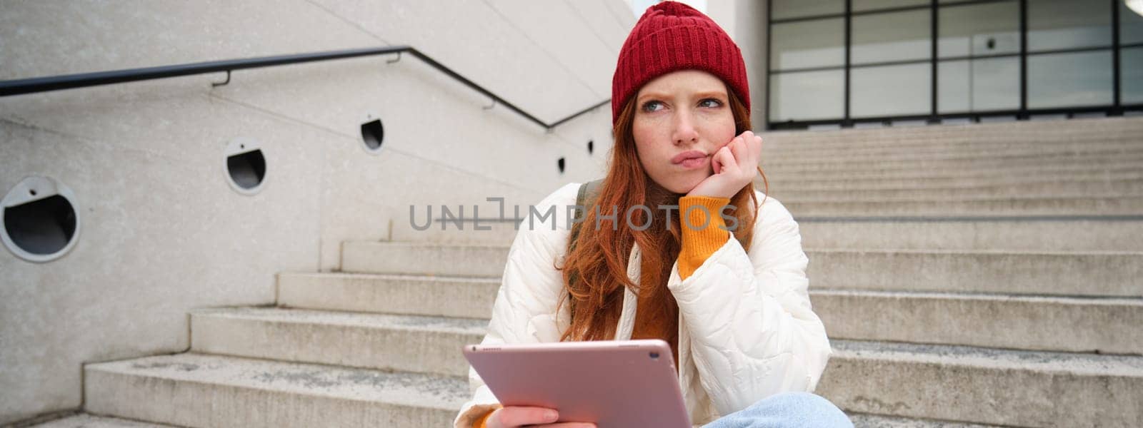 Portrait of troubled redhead girl, college student with thoughtful face, sits on street stairs, holds digital tablet, thinks how to reply on message by Benzoix