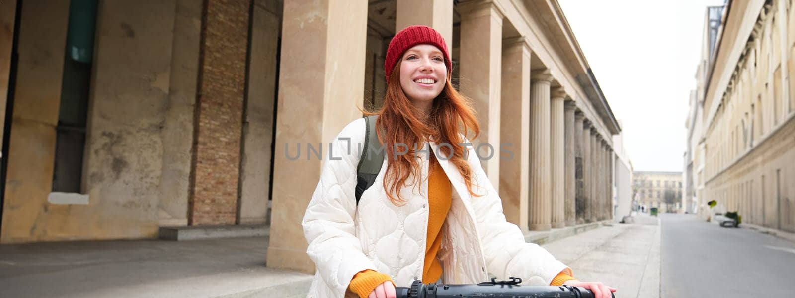 Young smiling redhead girl, student rides electric scooter, rents it and travels around city by Benzoix